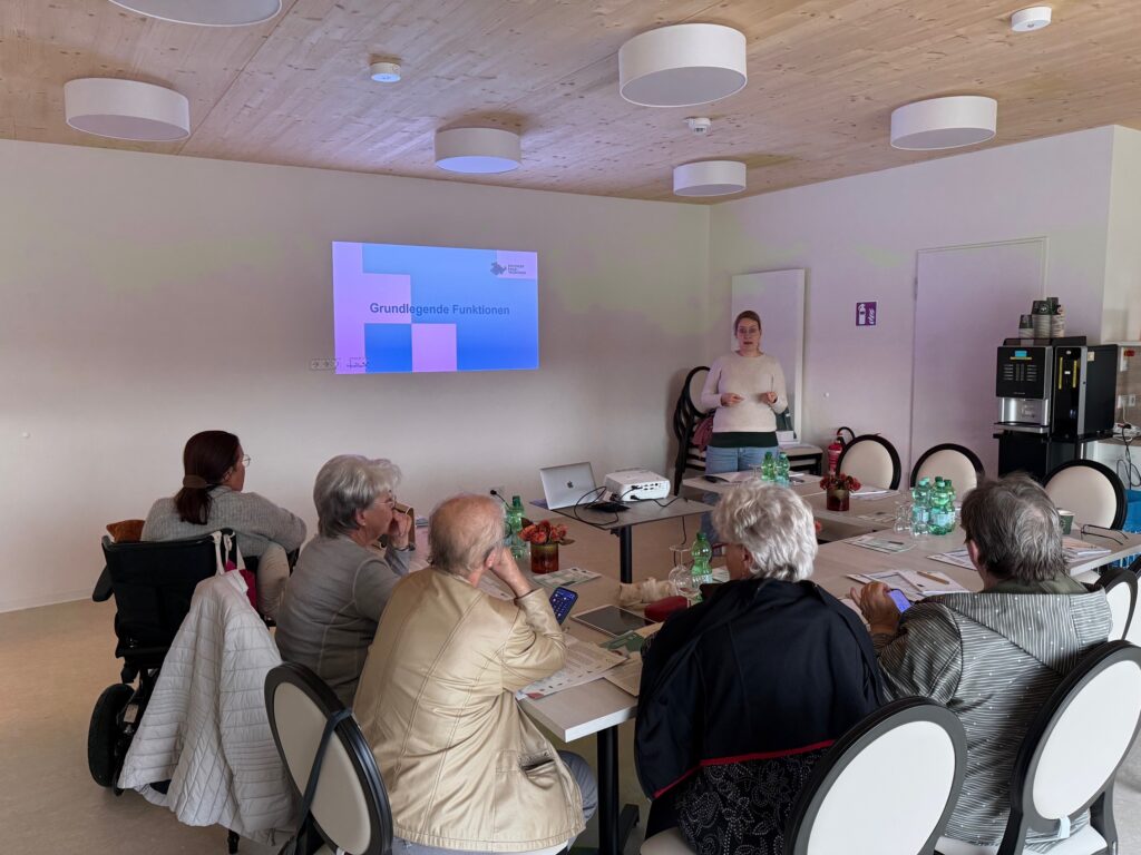 Ein Bild von der Veranstaltung in Isseroda. Senior:innen sitzen in einem Raum. An der Wand ist eine PowerPoint-Präsentation projeziert. Martina steht vor der Gruppe und redet.
