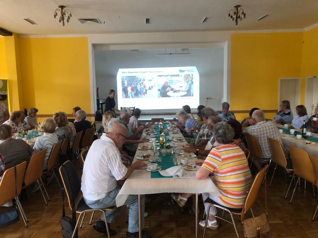 Das Bild zeigt eine größere Gruppe von Seniorinnen und Senioren, die an langen, gedeckten Tischen in einem gelb gestrichenen Raum sitzen und einer Präsentation des "Digitalen Engel Thüringen" folgen. Die Mobilreferentin steht vorne neben der Leinwand, auf der eine Präsentation mit Bildern und dem Titel "Der Digitale Engel Thüringen" angezeigt wird. Auf den Tischen sind Geschirr, Kaffee und Kuchen arrangiert, was auf eine gemütliche Atmosphäre hindeutet. Die Teilnehmer*innen scheinen aufmerksam zuzuhören und sind in eine lockere Gesprächsrunde eingebunden.