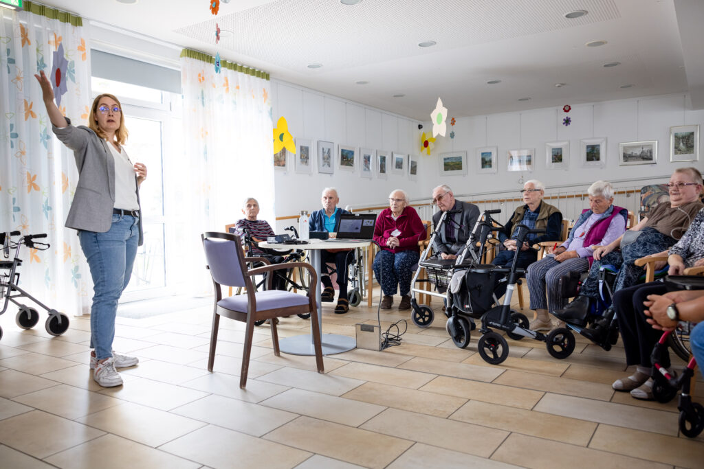 Eine Gruppe von Senioren hört einem Vortrag zu, während eine Referentin spricht. Der Raum ist hell und freundlich gestaltet.