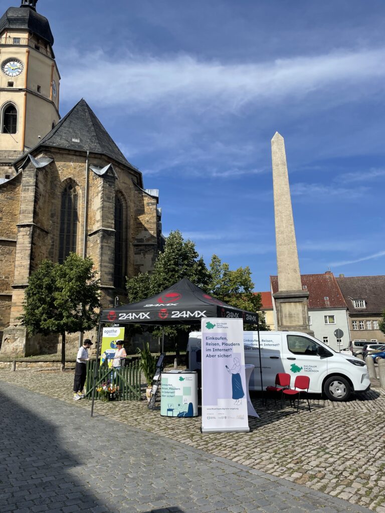 Informationsstand vom Digitalen Engel Thüringen mit einem Zelt, einem Fahrzeug und Werbematerialien vor einer Kirche und einem Obelisken.