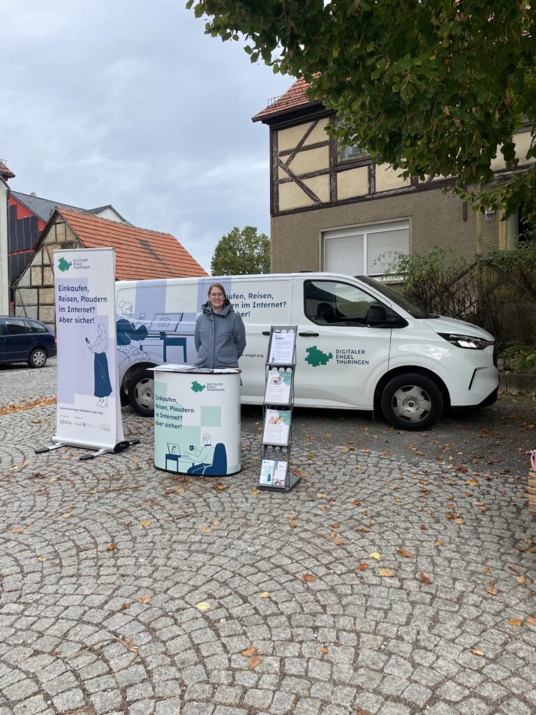 Das Bild zeigt das Infomobil des "Digitalen Engel Thüringen" auf einem gepflasterten Platz vor Fachwerkhäusern. Die Mobilreferentin steht neben dem Fahrzeug, das mit dem Slogan „Einkaufen, Reisen, Plaudern im Internet? Aber sicher!“ beschriftet ist. Vor dem Infomobil sind ein Roll-up-Banner, eine Infotheke und ein Prospektständer mit weiteren Informationsmaterialien aufgebaut. Die Bäume im Hintergrund lassen vermuten, dass das Bild im Herbst aufgenommen wurde, da einige Blätter bereits auf dem Boden liegen. Die Atmosphäre wirkt ruhig und informativ.