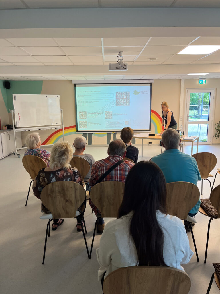 Das Bild zeigt eine kleine Gruppe älterer Personen, die in einem modernen Seminarraum auf Holzstühlen sitzen und einer Präsentation der Mobilreferentin des "Digitalen Engel Thüringen" folgen. Die Referentin steht vorne neben der Leinwand und präsentiert Informationen, die unter anderem QR-Codes enthalten. Im Raum befinden sich links eine Tafel und ein Deckenprojektor. Die farbenfrohe Wandbemalung mit Regenbogenstreifen verleiht dem Raum eine freundliche und einladende Atmosphäre.