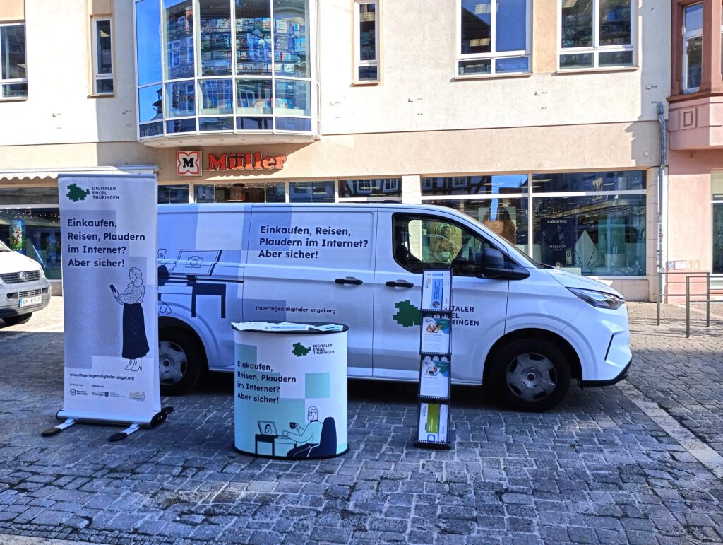 Das Bild zeigt das Infomobil des "Digitalen Engel Thüringen", einen weißen Transporter mit der Aufschrift und dem Slogan "Einkaufen, Reisen, Plaudern im Internet? Aber sicher!", der auf einem Platz vor einem Müller-Drogeriemarkt geparkt ist. Neben dem Fahrzeug steht ein Roll-up-Banner mit dem gleichen Slogan und einer Illustration einer Person mit einem Smartphone. Ein kleiner Infostand sowie ein Prospektständer mit Flyern ergänzen das Setup.