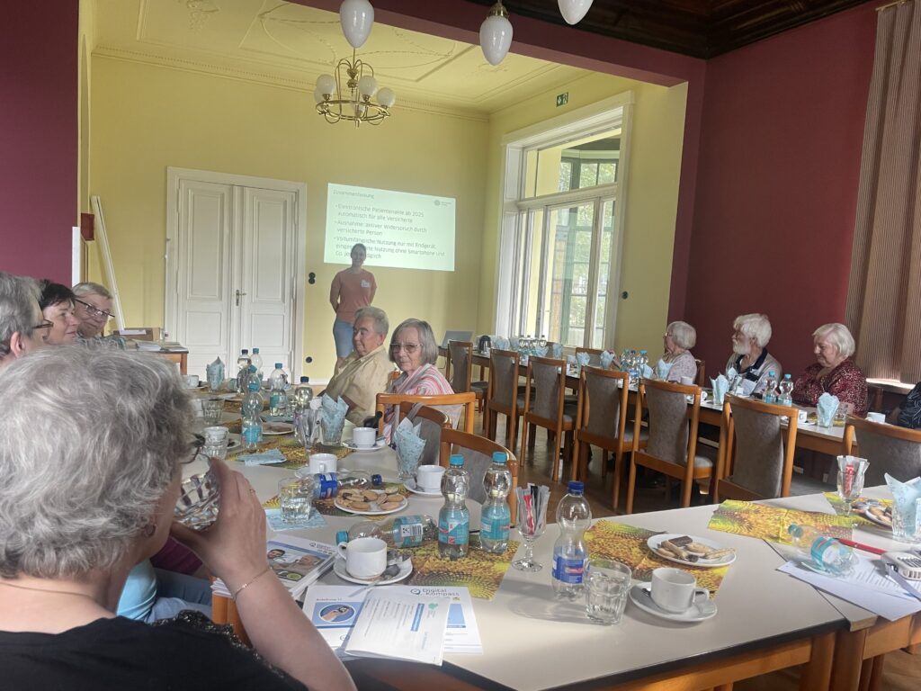 Das Bild zeigt eine Gruppe älterer Personen, die in einem gemütlichen Raum an gedeckten Tischen sitzen und einer Präsentation folgen. Eine Referentin steht vorne neben einer Leinwand, auf der eine Folie mit dem Titel "Zusammenfassung" und einigen Stichpunkten angezeigt wird. Auf den Tischen sind Wasserflaschen, Gläser, Tassen und kleine Teller mit Gebäck verteilt. Die Wände des Raumes sind in warmen Gelb- und Rottönen gehalten, und ein Kronleuchter hängt von der Decke, was dem Raum eine einladende Atmosphäre verleiht.