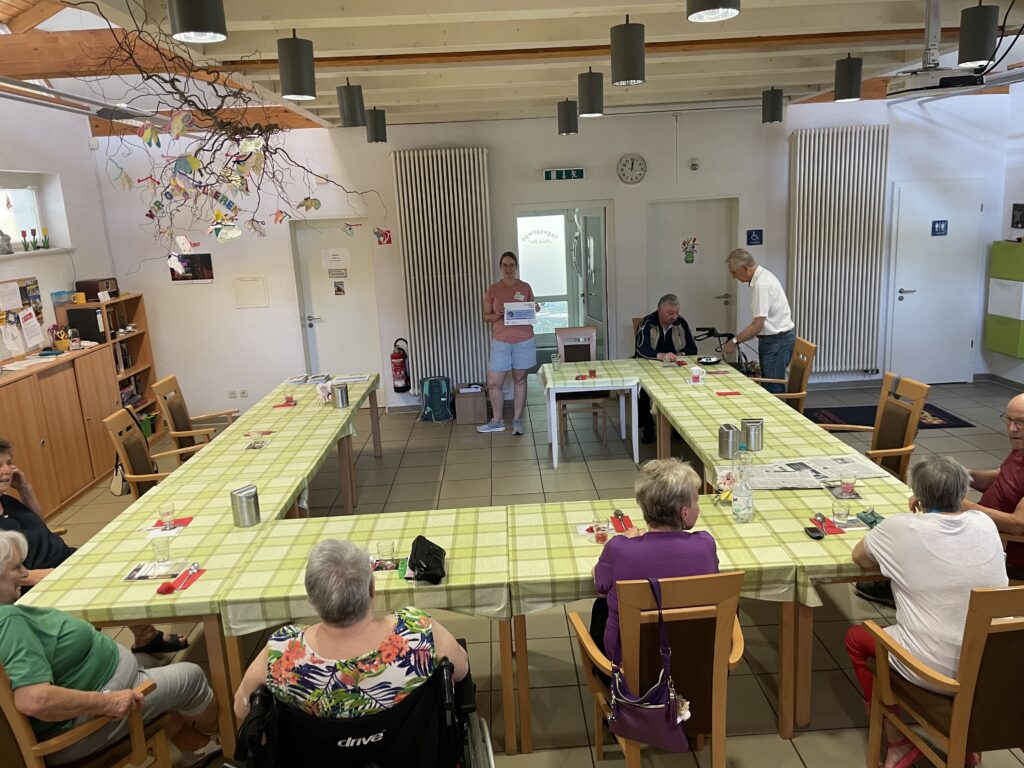 Das Bild zeigt eine Gruppe älterer Menschen, die in einem Gemeinschaftsraum an Tischen sitzen, die in einer offenen U-Form angeordnet sind. Eine Referentin steht am Ende der Tische und hält ein Präsentationsmaterial in den Händen, während ein Mann mit einem Rollator daneben steht. Der Raum ist freundlich eingerichtet, mit einer grünen Tischdecke und einer hängenden Dekoration aus bemalten Papierfischen und Zweigen an der Decke. Im Hintergrund sind verschiedene Türen und ein Schrank mit Büchern und Dekoration zu sehen, was auf eine einladende und kreative Atmosphäre hinweist.