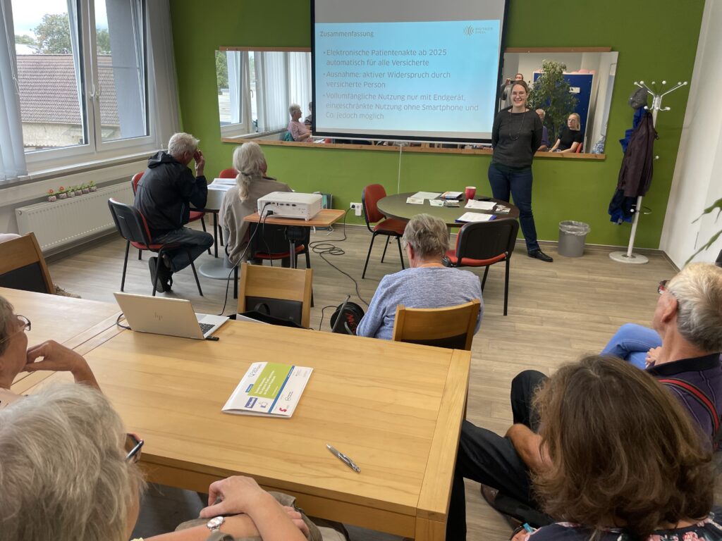 Das Bild zeigt eine Präsentationsveranstaltung des "Digitalen Engel Thüringen" in einem Mehrgenerationenhaus. Eine Referentin steht vor einer Leinwand und präsentiert Inhalte zur elektronischen Patientenakte, die ab 2025 für alle Versicherten eingeführt werden soll. Die Teilnehmenden, überwiegend ältere Menschen, sitzen in lockerer Runde und hören aufmerksam zu. Vor ihnen auf dem Tisch liegen Informationsmaterialien und ein Laptop ist ebenfalls aufgeklappt, vermutlich zur Unterstützung der Präsentation. Der Raum ist freundlich gestaltet, mit grünen Wänden und großen Fenstern, die viel Tageslicht hereinlassen. Ein Spiegel an der Wand reflektiert einen Teil der Gruppe, was den Raum optisch erweitert.