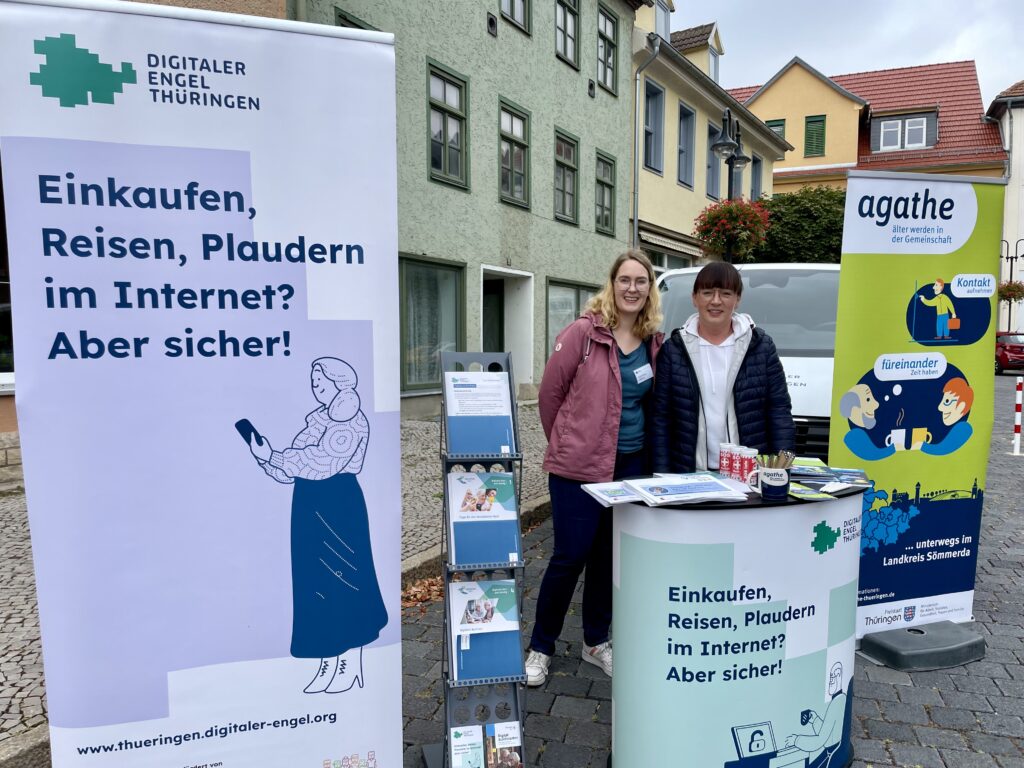 Das Bild zeigt zwei Frauen, die an einem Infostand des "Digitalen Engel Thüringen" auf einem Marktplatz stehen. Links ist ein großes Roll-up-Banner mit dem Slogan „Einkaufen, Reisen, Plaudern im Internet? Aber sicher!“ und einer Illustration einer älteren Person mit Smartphone zu sehen. Neben dem Stand ist ein weiteres Banner von „agathe – älter werden in der Gemeinschaft“, das für ein Projekt im Landkreis Sömmerda wirbt. Auf dem Tisch liegen Informationsmaterialien und Broschüren aus. Im Hintergrund sind bunte Häuserfassaden und das Infomobil des "Digitalen Engel Thüringen" erkennbar.