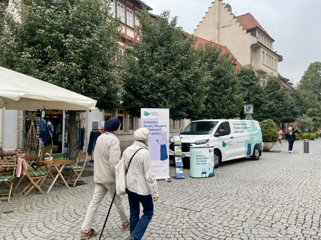 Das Bild zeigt das Infomobil des "Digitalen Engel Thüringen" auf einem gepflasterten Marktplatz in Bad Langensalza. Vor dem weißen Transporter, der den Slogan „Einkaufen, Reisen, Plaudern im Internet? Aber sicher!“ trägt, stehen ein Roll-up-Banner und eine Infotheke mit weiteren Informationsmaterialien. Links im Bild gehen zwei ältere Personen an dem Stand vorbei, während rechts eine Person in den Hintergrund spaziert. Im Umfeld sind Bäume, ein Café mit Außenbestuhlung und historische Gebäude mit verzierten Fassaden zu sehen, was der Szene eine einladende, städtische Atmosphäre verleiht.