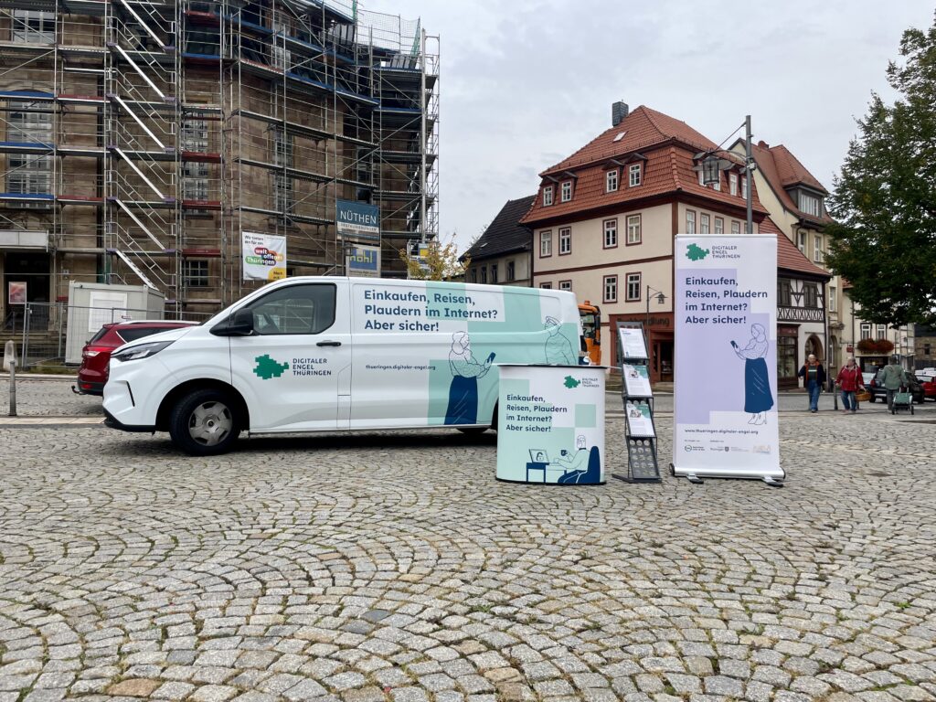 Das Bild zeigt das Infomobil des "Digitalen Engel Thüringen" auf einem gepflasterten Marktplatz in Waltershausen. Der weiße Transporter trägt den Slogan „Einkaufen, Reisen, Plaudern im Internet? Aber sicher!“ und ist zusammen mit einem Roll-up-Banner, einer Infotheke und einem Prospektständer aufgestellt. Im Hintergrund befinden sich historische Gebäude mit traditionellen Fassaden sowie ein Gerüst an einem renovierten Bauwerk, was auf laufende Sanierungsarbeiten hinweist. Einige Passanten sind am Rande des Platzes unterwegs, was der Szenerie eine lebendige, städtische Atmosphäre verleiht.