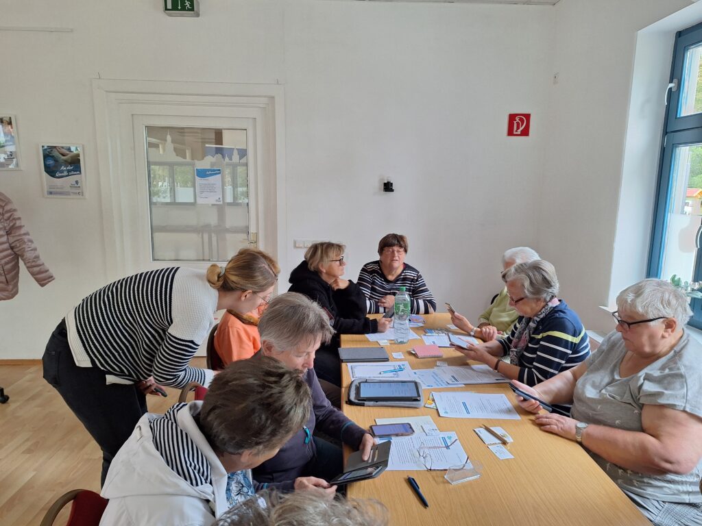 Das Bild zeigt eine Gruppe älterer Personen, die um einen Tisch versammelt sind und gemeinsam mit der Mobilreferentin des "Digitalen Engel Thüringen" an ihren Smartphones arbeiten. Die Mobilreferentin beugt sich zu einer Teilnehmerin und gibt individuelle Hilfestellung. Auf dem Tisch liegen mehrere Informationsblätter, Stifte und eine Wasserflasche. Der Raum ist schlicht gestaltet, mit weißen Wänden und großen Fenstern, die natürliches Licht hereinlassen. Die Atmosphäre wirkt konzentriert, da die Teilnehmer*innen offenbar etwas über die Nutzung digitaler Geräte lernen.