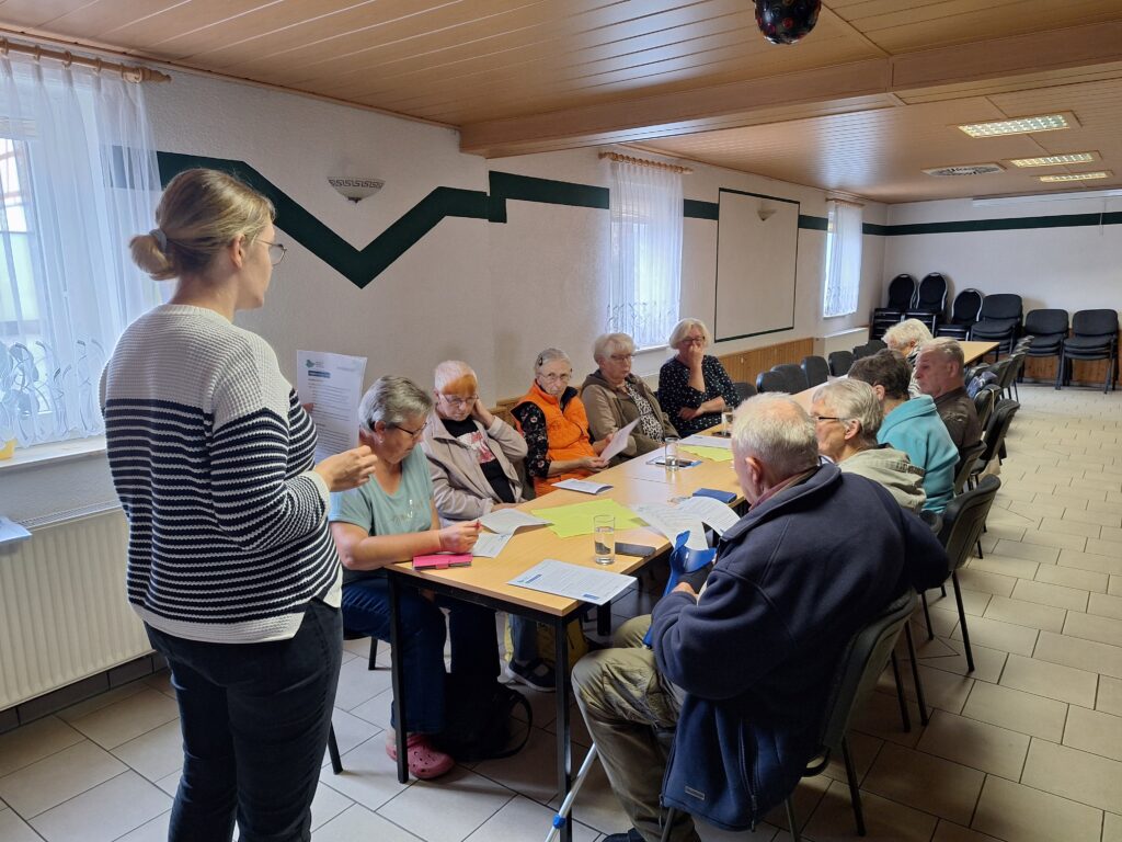 Das Bild zeigt die Mobilreferentin des "Digitalen Engel Thüringen", die vor einer Gruppe älterer Menschen steht und eine Schulung leitet. Die Teilnehmenden sitzen an einem langen Tisch und haben Informationsblätter und Notizen vor sich liegen. Die Mobilreferentin hält ein Dokument in der Hand und erklärt offenbar Inhalte, während die Teilnehmer:innen aufmerksam zuhören. Der Raum ist schlicht eingerichtet, mit weißen Wänden, Holzelementen an der Decke und gestapelten Stühlen im Hintergrund. Die Atmosphäre wirkt konzentriert und lehrreich.