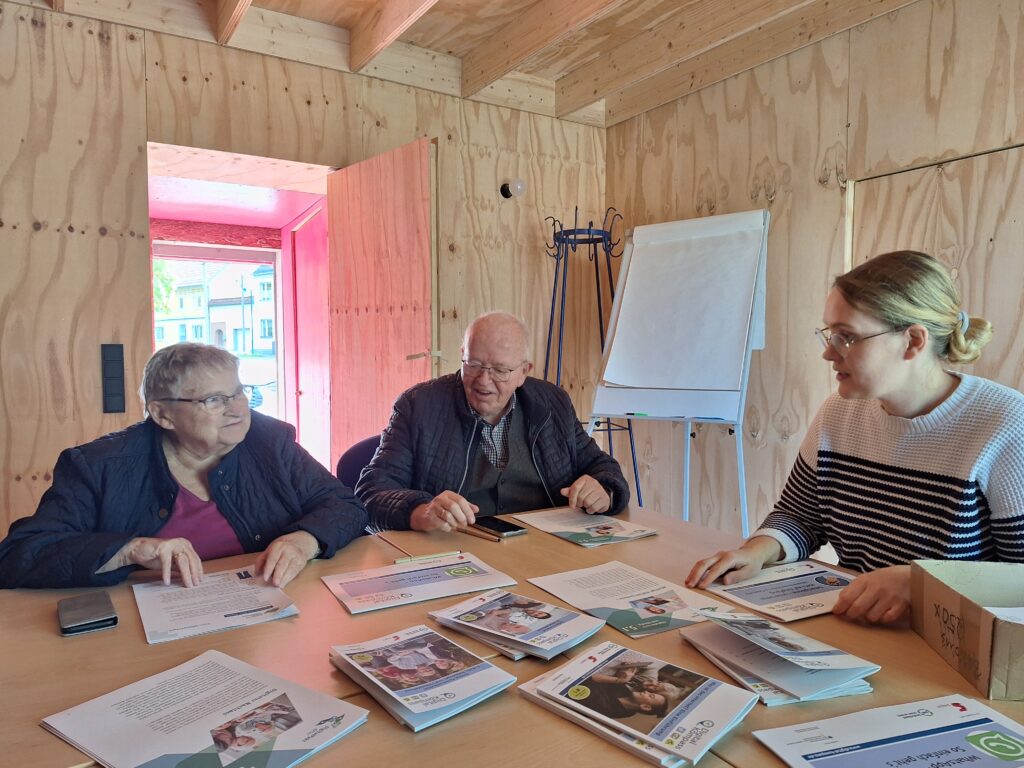 Das Bild zeigt drei Personen, zwei ältere Menschen und die Mobilreferentin des "Digitalen Engel Thüringen", die an einem Holztisch in einem Raum mit unbehandelten Holzverkleidungen sitzen. Auf dem Tisch liegen verschiedene Informationsbroschüren und Dokumente, die sie gemeinsam durchsehen. Die Mobilreferentin erklärt den beiden anderen etwas aus den Unterlagen. Im Hintergrund befinden sich ein Flipchart und eine Garderobe. Durch die offene Tür fällt Tageslicht in den Raum und beleuchtet die Szene.