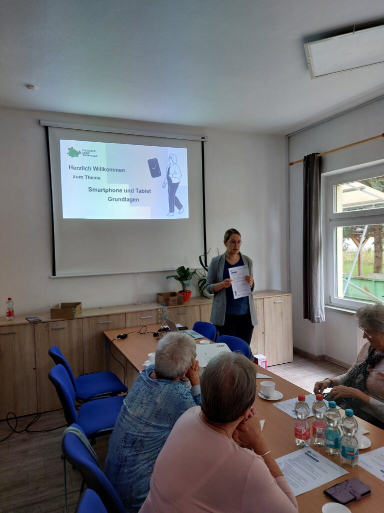 Martina steht vor einer Präsentation, die an die Wand geworfen wird. Vor ihr sitzt eine Gruppe älterer Menschen