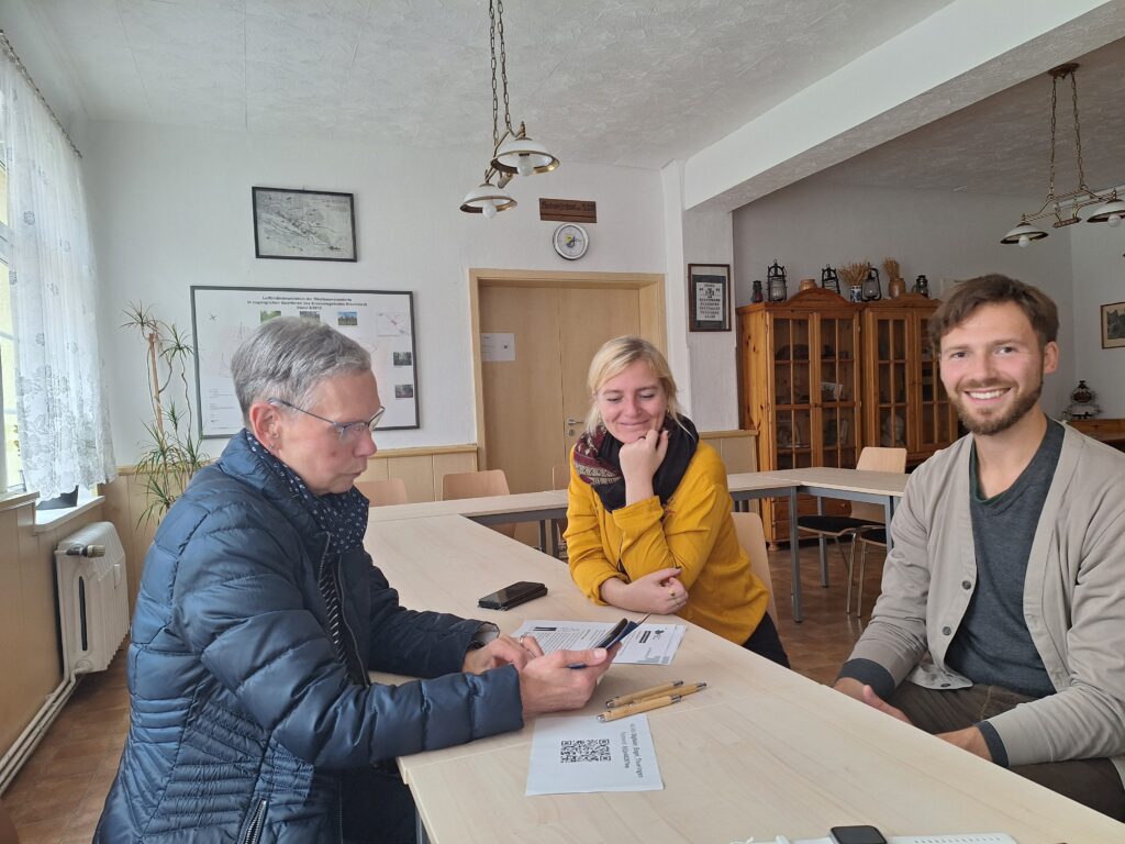 Martina, Timo und eine ältere Frau sitzen an einem Tisch. Die Frau hält ein Handy in der Hand.