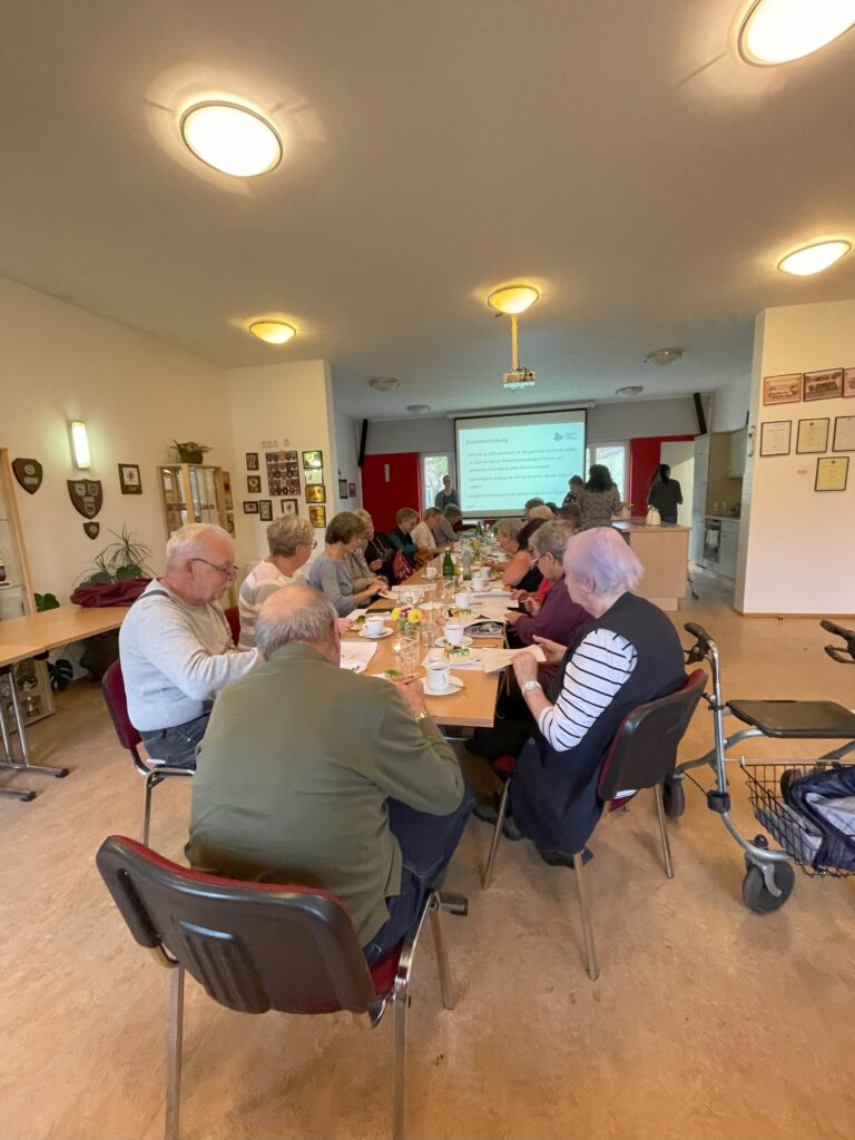 Das Bild zeigt eine Gruppe älterer Teilnehmer:innen, die an einem langen Tisch in einem Gemeindesaal sitzen und einer Präsentation folgen. Auf dem Tisch stehen Geschirr, Gläser und Notizmaterialien, was auf eine gemütliche Veranstaltung mit Kaffee und Diskussionen hindeutet. Im Hintergrund ist eine Leinwand zu sehen, auf der die Mobilreferentin des "Digitalen Engel Thüringen" Inhalte präsentiert. Der Raum ist schlicht gestaltet, mit weißen Wänden, roten Akzenten und dekorativen Schildern an der Wand. Ein Rollator steht am Rand des Raumes, was auf eine barrierefreie Umgebung hinweist.
