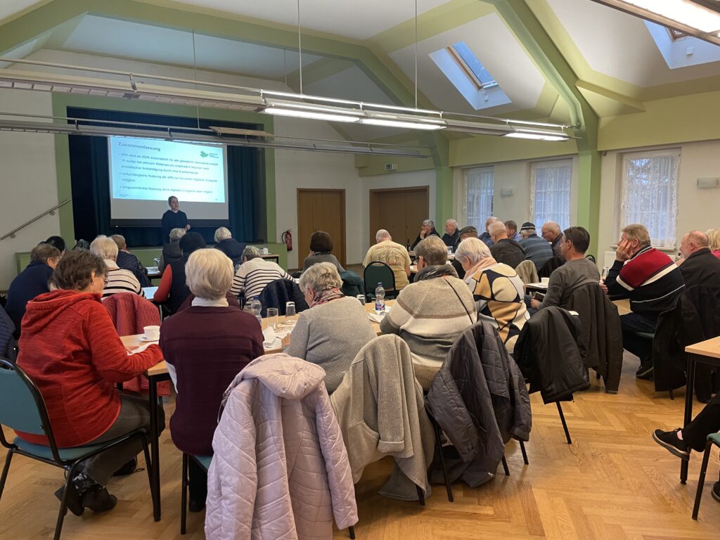 Das Bild zeigt eine größere Gruppe älterer Teilnehmer:innen, die in einem Saal an Tischen sitzen und einer Präsentation folgen. Vorne steht die Mobilreferentin des "Digitalen Engel Thüringen" und spricht vor einer Leinwand, auf der eine Folie mit dem Titel "Zusammenfassung" zu sehen ist. Der Saal ist hell und geräumig, mit grünen Wänden, hohen Decken und Fenstern mit weißen Vorhängen. Auf den Tischen befinden sich Kaffeetassen und Wassergläser, was auf eine gemütliche und informative Veranstaltung hinweist. Die Teilnehmenden wirken aufmerksam und konzentriert.