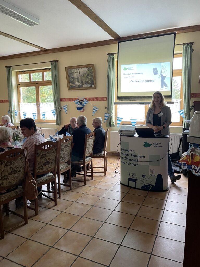 Martina steht hinter einem Rednerpult. Hinter ihr ist die Präsentation an eine Leinwand projiziert. Ältere Menschen sind im Rest des Raums zu sehen.