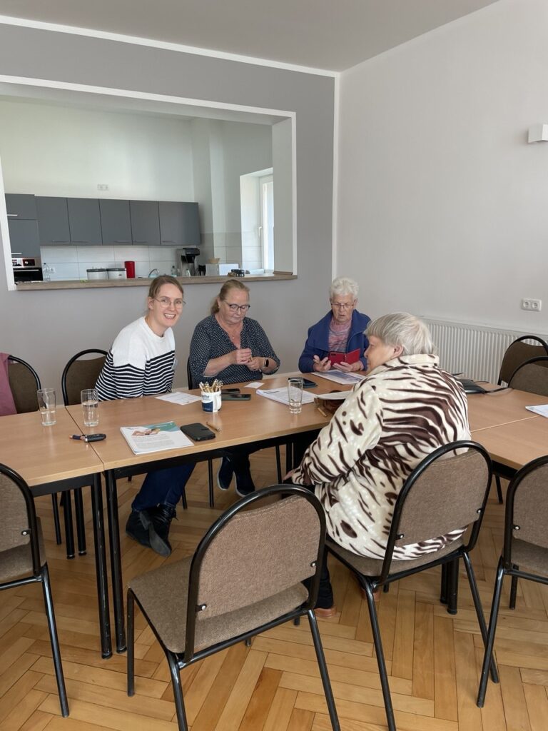 Martina sitzt mit meherern älteren Menschen an einem Tisch. Informationsmaterialien liegen auf dem Tisch.