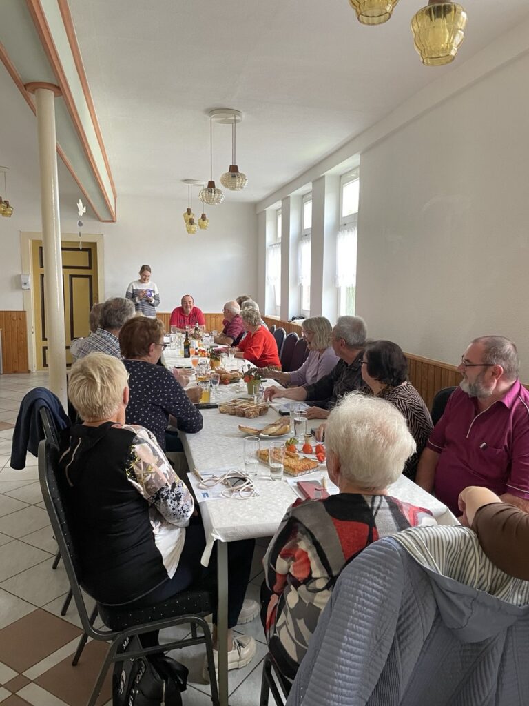 Eine große Tafel mit vielen älteren Menschen. Ganz vorne sieht man Martina, die zu ihnen spricht.