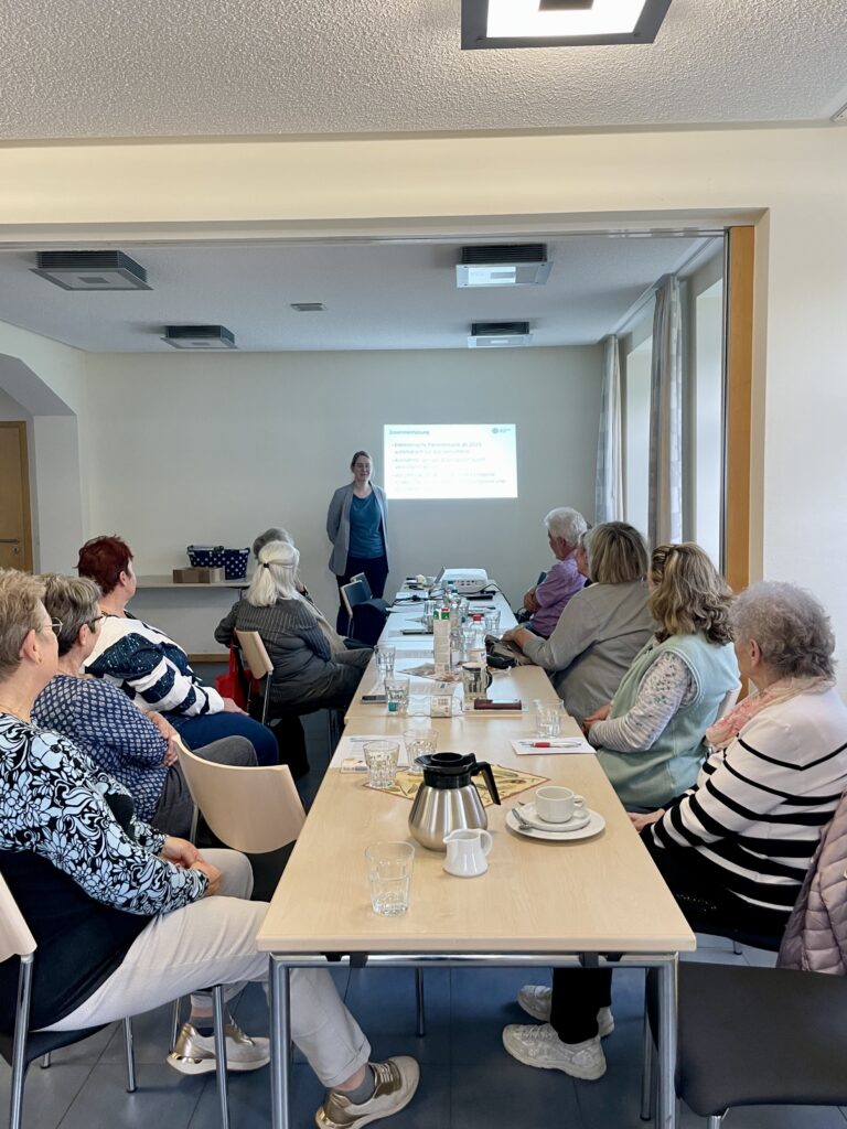 Eine lange Tafel an der ältere Menschen sitzen. Am Ende der Tafel ist Martina zu sehen. Hinter ihr ist eine Präsentation an die Wand geworfen