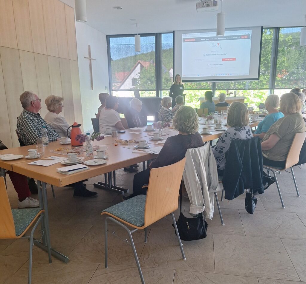 Martina steht vor einer Gruppe sitzender Senior:innen in einem Raum mit großen Fenstern.