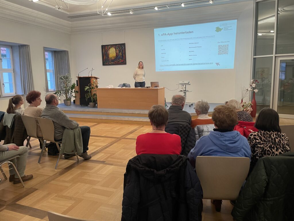 Martina steht vor einer Gruppe ältere Menschen in einem großen Saal. Hinter Martina ist eine Präsentation an die Wand projiziert.