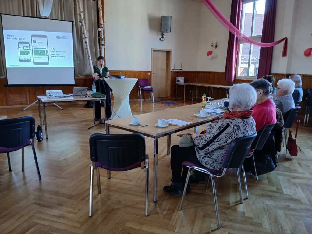Ein Saal in dem vorne im Bild ältere Menschen an einem Tisch sitzen. Im Hintergrund steht die Mobilreferentin Sophie vor einer Leinwand, auf die eine Präsentation projiziert wird.