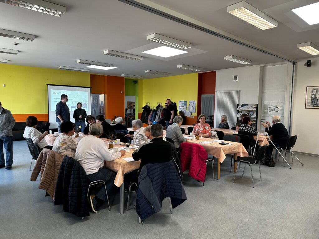 Ein Raum mit einer großen Sitzgruppe. Hinten im Bild ist Sophie vor einer Leinwand zu sehen. Sie spricht zu den anwesenden Senior:innen.