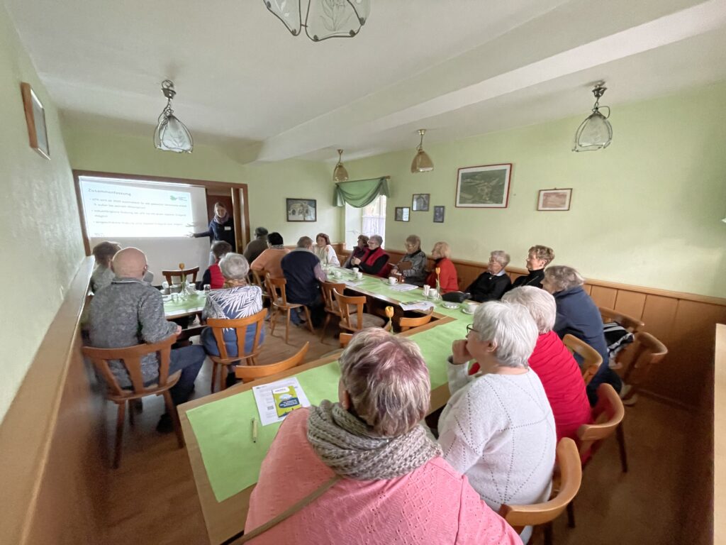 Ältere Menschen sitzen in einem großen Raum. Am Ende des Raumes steht Martina vor ihrer Präsentation.
