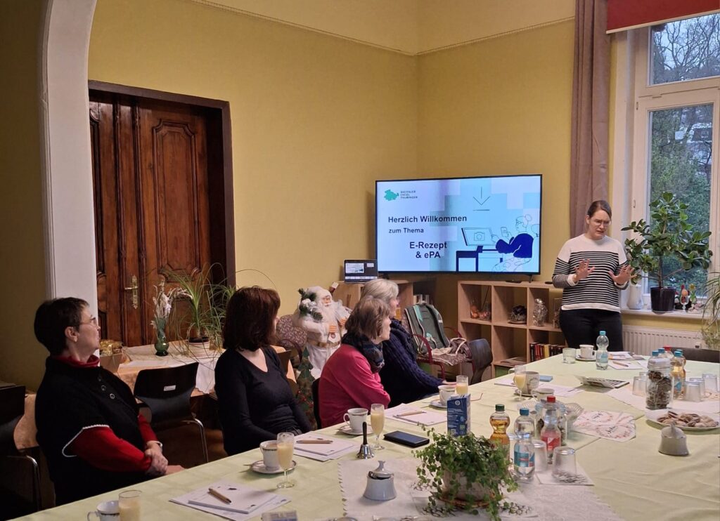 Martina steht vor einer Gruppe Senior:innen in einem Raum. Sie steht vor einem Bildschirm