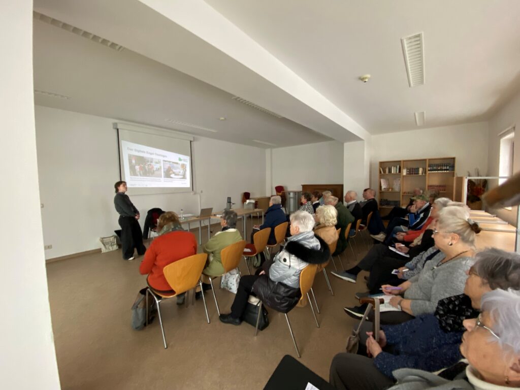 Ein Raum, in dem mehrere ältere Menschen sitzen. Ganz vorne im Raum steht Sophie. Hinter ihr ist eine Präsentation an die Wand projiziert.