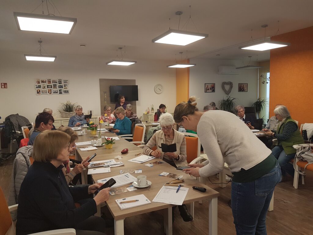 Martina hilft einer Person mit ihrem Smartphone. Sie beugt sich über einen Tisch, an dem mehrere Personen sitzen.