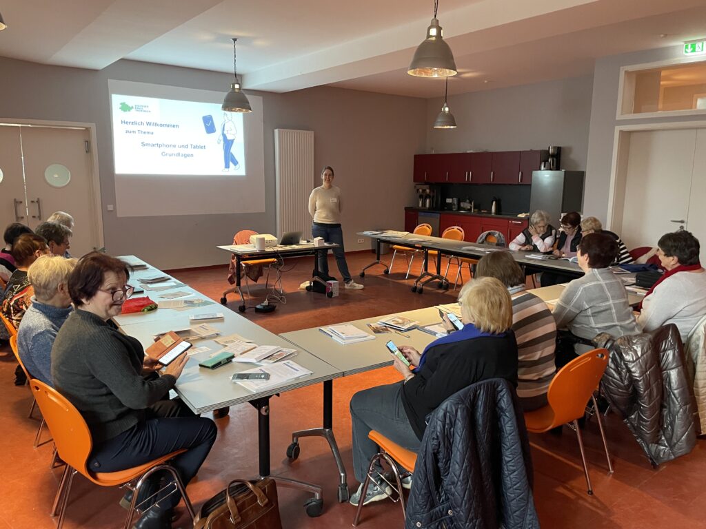 Ein Raum mit einer Sitzgruppe an der ältere Menschen sitzen. In der Mitte steh Martina und spricht zu den Personen. Ab die Wand ist eine Präsentation projiziert.
