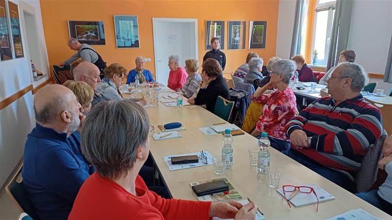 Ein gut gefüllter Raum mit einer langen Tafel. Am Ende des Tisches steh Sophie und spricht zu den Anwesenden.