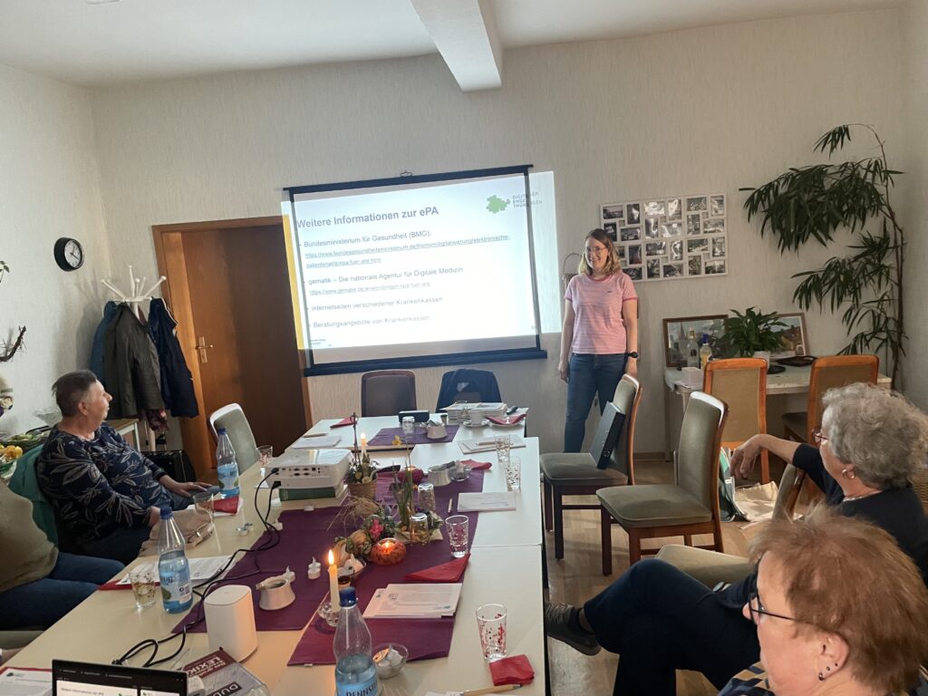 Ein kleiner Raum mit einem großen Tisch und einer Leinwand, an die eine Präsentation projiziert ist.