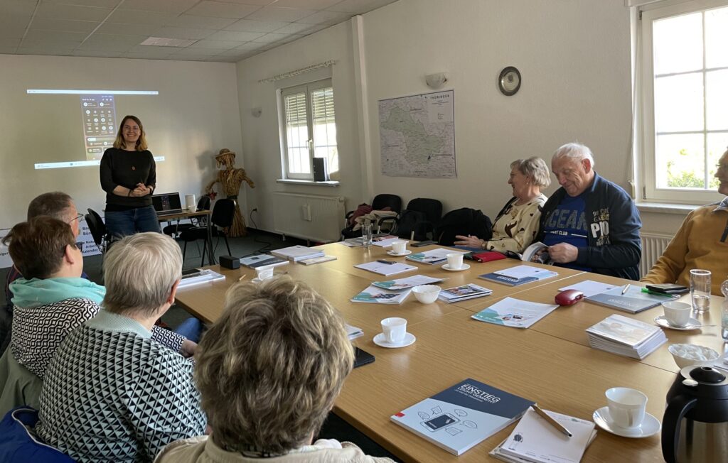 Ein Raum mit einem großen Tisch an dem ältere Menschen sitzen. Am Ende des Tisches steht Martina und spricht zur Gruppe.