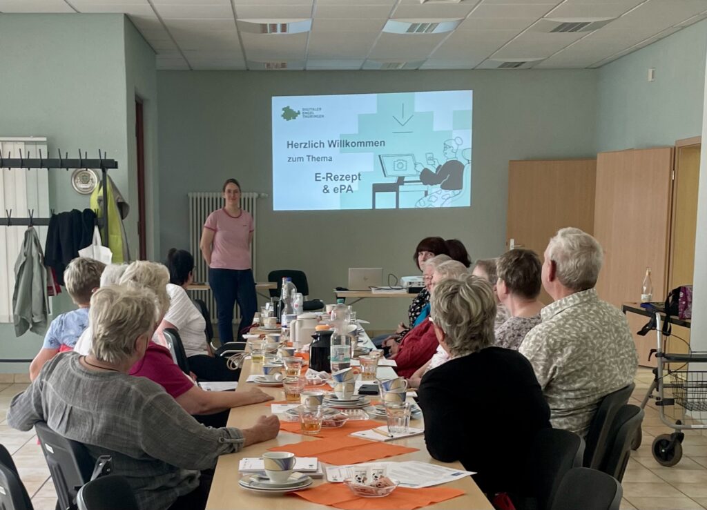 Martina steht lächelnd vor einer Gruppe ältere Menschen, die an einem großen TIsch sitzen. Hinter ihr ist eine Präsentation auf die Wand projiziert.