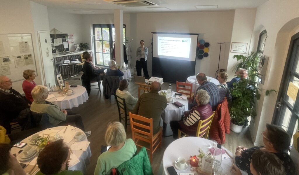 Martina steht vor einer Gruppe Senior:innen in der Begegnungsstätte und hält einen Vortrag,