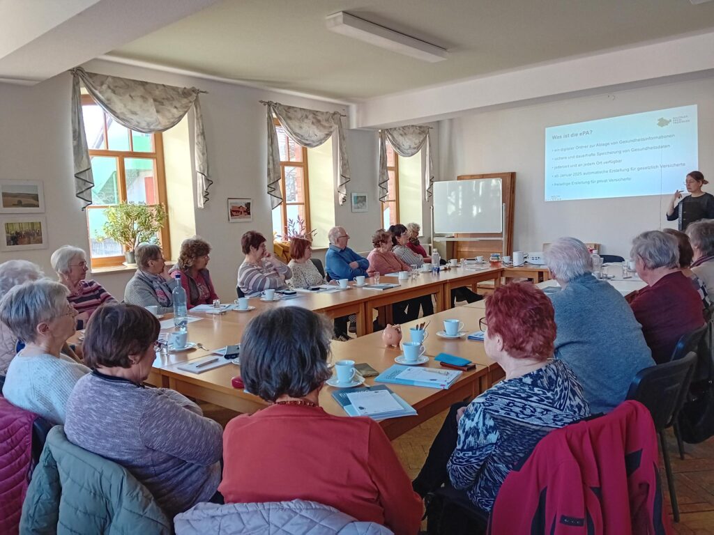 Ein Raum mit mehreren langen Tischen gefüllt, an denen ältere Menschen sitzen. Am Ende des Raumes ist Martina und spricht zu den Anwesenden.
