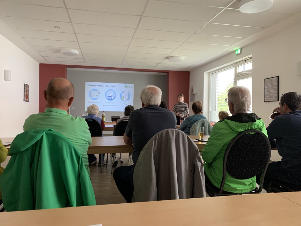 Sophie steht vor einer Gruppe sitzender Senior:innen in einem Saal. Hinter ihr ist eine Präsentation an die Wand geworfen und sie spricht zur Gruppe.