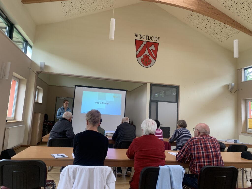 Eine Gruppe älterer Menschen in einem Raum in der Rückenansicht. Ganz vorne steht Martina im Raum und spricht zu ihnen.