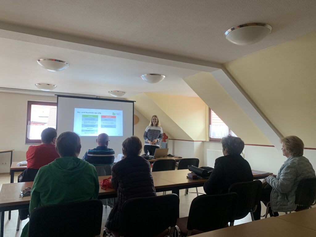 Ein Raum mit einem angeschrägten Dach. Martina spricht zu einer Gruppe älterer Menschen.