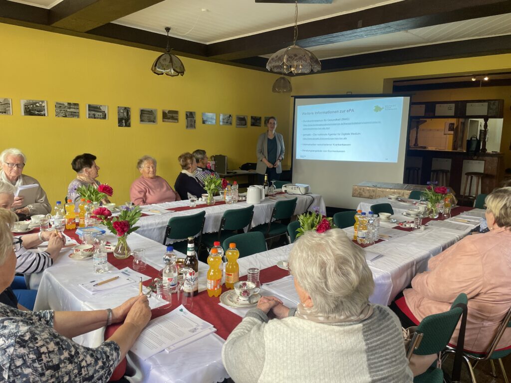 Martina steht für einer Gruppe älterer Menschen, die an einer Tischrunde sitzen. Hinter Martina ist eine Präsentation an die Wand projiziert.
