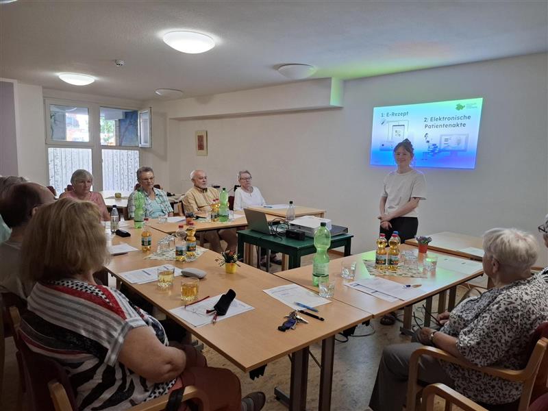 Eine große Sitzgruppe in U-Form. Vorne steht Sophie und schaut freundlich in Richtung Kamera. Hinter ihr ist eine Präsentation projiziert.