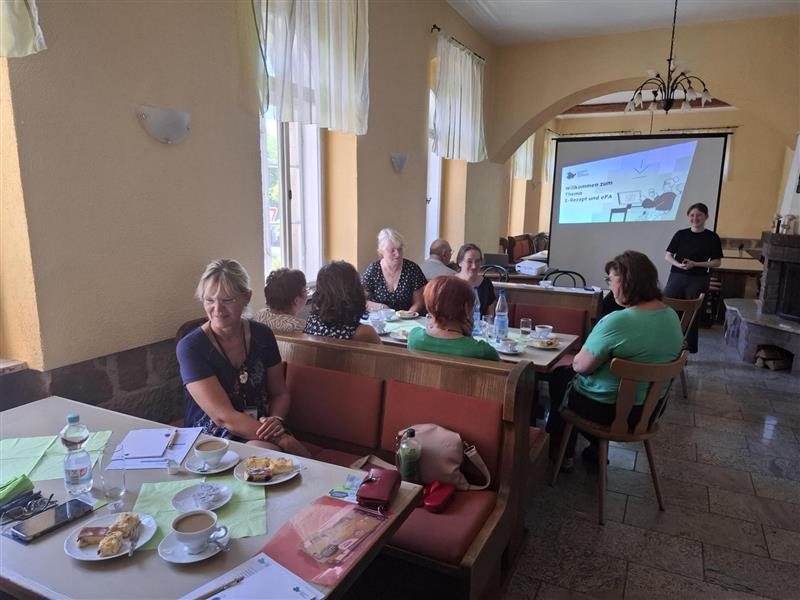 Ein Raum, in dem Menschen in Sitzgruppen zusammensitzen. Getränke und Essen stehen auf den Tischen und alle sehen sehr freundlich ausgelassen aus. Sophie steht vor der Gruppe und hält einen Vortreag.