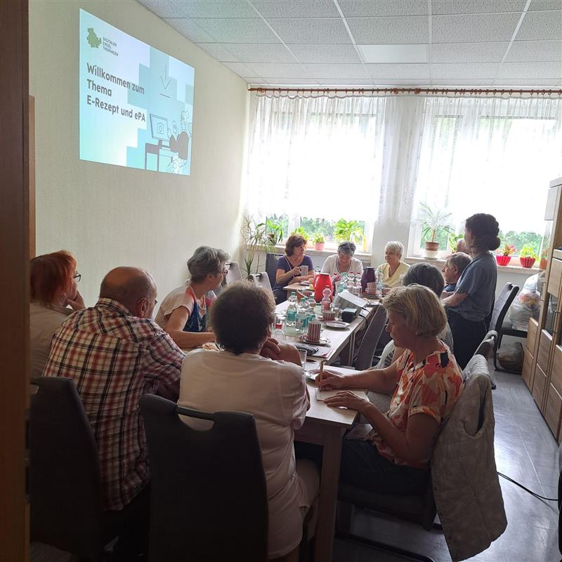 Sophie steht hinter einer Gruppe älterer Menschen, die an einem Tisch sitzt. Sie spricht mit ihnen. An der Wand vor ihnen ist eine Präsentation an die Wand projiziert.