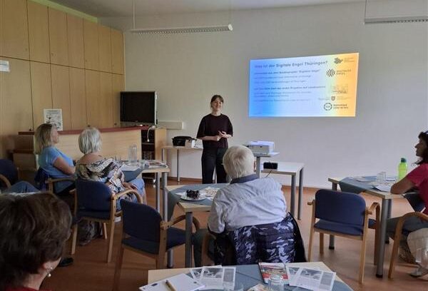 Sophie steht vor einer Gruppe älterer Menschen, die an Tischen sitzt. Auf den Tischen sind Informationsmaterialien verteilt.