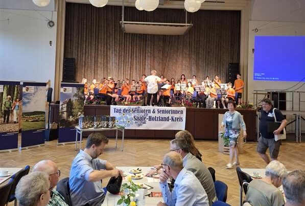 Es ist eine Bühne zu sehen. Auf der Bühne ist ein Chor und vor der Bühne sind Tische uafgebaut, an denen Menschen sitzen.