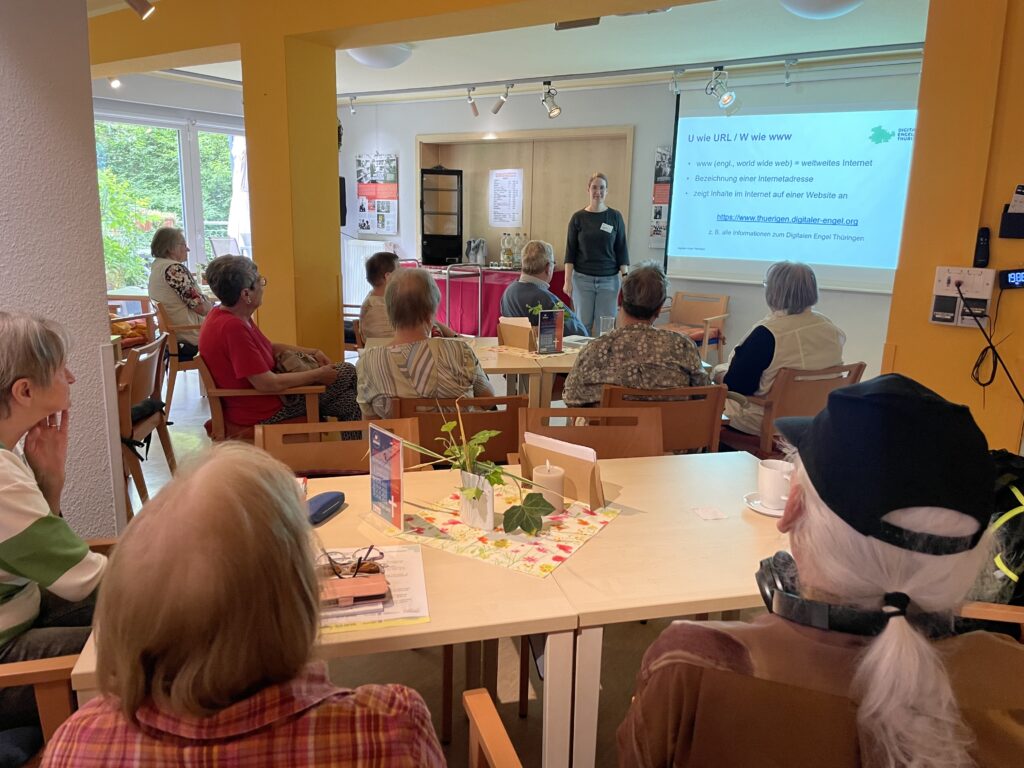 Ein großer gefüllter Raum. Vorne steht Martina und lächelt in die Kamera.