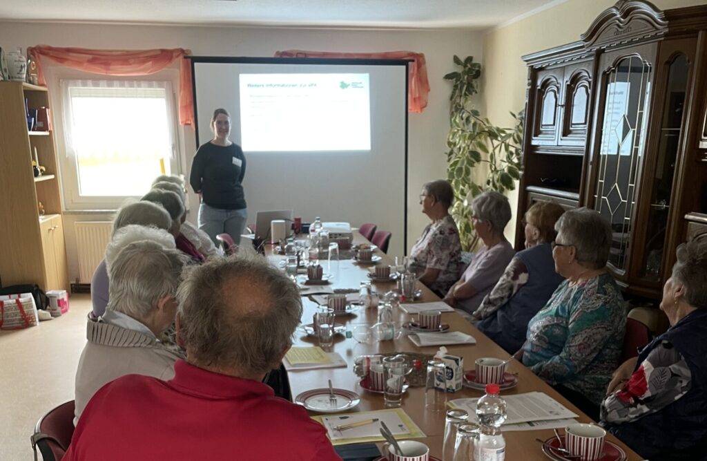 Martina steht vor einer Gruppe SEnior:innen, die an einem langen Tisch sitzen.